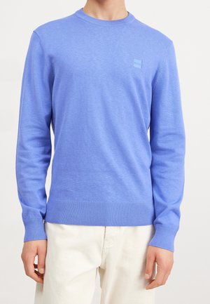 Homme portant un pull bleu clair à col rond manches longues avec un petit logo sur la poitrine et un pantalon couleur crème.