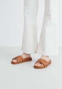 Sandali a scivolo marroni, con un design della fascia intrecciata, suola piatta in gomma e punta aperta. Indossati con pantaloni bianchi a gamba larga su uno sfondo chiaro.