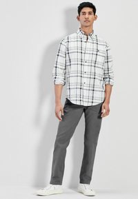Camisa a cuadros blanca y negra con cierre de botones, combinada con pantalones grises y zapatillas blancas. El modelo se encuentra de pie frente a un fondo liso.