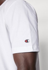 T-shirt blanc en coton avec un col rond, arborant un petit logo brodé en rouge et noir sur la manche. Texture lisse, manches courtes.