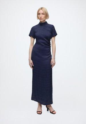 Femme aux cheveux blonds courts portant une robe maxi bleu marine ajustée à manches courtes et des talons ouverts noirs, debout devant un fond blanc uni.