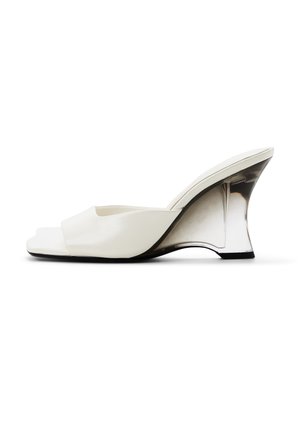 Mule blanco de punta abierta con una tira ancha y un tacón cuña alto, curvado y transparente, sobre un fondo blanco.