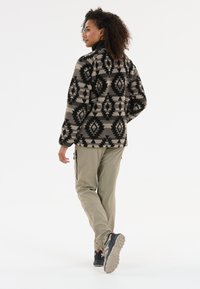 Veste en polaire avec un motif géométrique noir et beige, col montant et manches longues, associée à un pantalon cargo beige clair et des chaussures robustes.