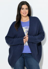 Mörkblå oversized stickad kofta med långa ärmar och en vid öppen front. Strukturen är ribbad med en mjuk finish. Bärs över en grafisk t-shirt.