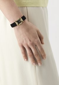 Pols met zwart leren horloge met een gouden, van edelstenen bezet rechthoekig gezicht dat rust tegen geplooide witte stof.