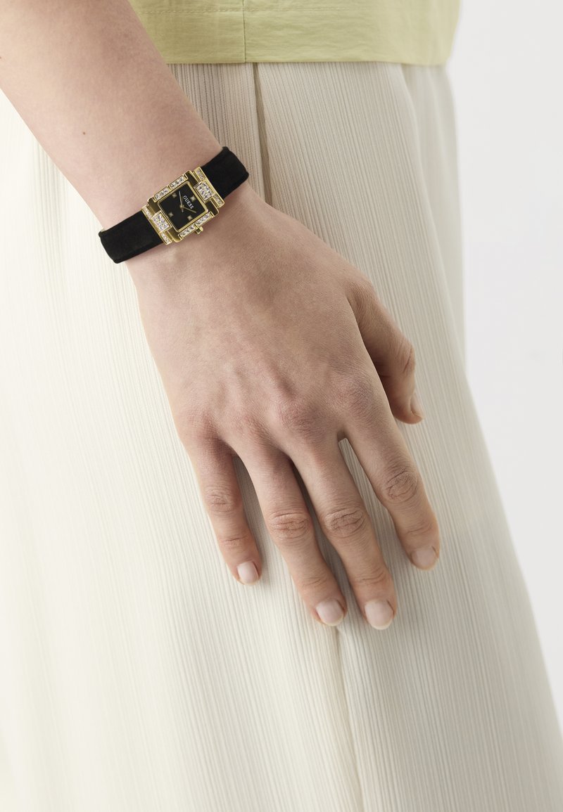 Pols met zwart leren horloge met een gouden, van edelstenen bezet rechthoekig gezicht dat rust tegen geplooide witte stof.