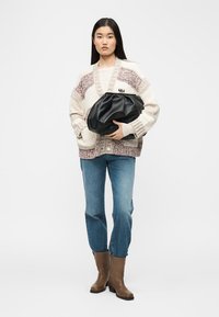 Γυναίκα που φοράει μπεζ και καφέ πλεκτή ζακέτα, μπλε τζιν, καφέ μπότες, κρατώντας μια μεγάλη μαύρη δερμάτινη τσάντα φάκελο σε λευκό φόντο.