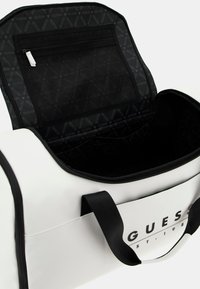 White duffel bag with black accents, featuring a zippered top, mesh pocket inside, and logo on the front. Smooth synthetic material.