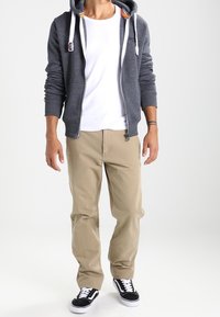 Sudadera gris con cremallera y cordones blancos, llevada sobre una camiseta blanca; los pantalones beiges y las zapatillas en blanco y negro completan el conjunto.