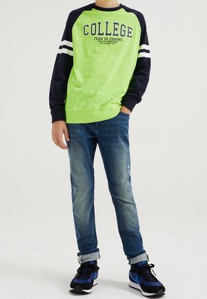 Zielona i granatowa bluza z napisem "COLLEGE" i paskami, zestawiona z niebieskimi dżinsami oraz czarnymi trampkami z niebieskimi akcentami.
