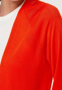 Jasno pomarańczowy kardigan o miękkiej fakturze, z otwartym przodem i misternym wzorem dzianiny na rękawach. Lekki i casualowy design.