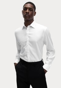 Camisa blanca de manga larga con cuello de botón y tapeta frontal, hecha de un tejido suave, combinada con pantalones negros.