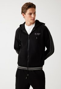 Felpa nera con zip, polsini e vita a coste, con un logo e motivi decorativi sulle maniche. Realizzata in materiale morbido e elasticizzato.