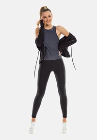 Sportoutfit met een grijs mouwloos shirt, zwarte strakke legging en een zwarte jas met ritssluiting en geplooide mouwen. Witte sportschoenen.