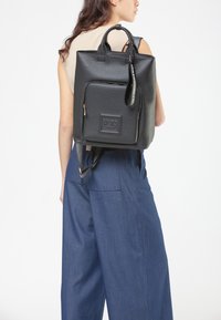 Donna che indossa pantaloni ampi blu e una maglietta beige senza maniche, che porta uno zaino in pelle nera testurizzata con tasca frontale con zip e manici superiori.