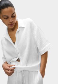 Camicia bianca a maniche corte, con texture e colletto, legata in vita. Abbinata a pantaloni leggeri con cordino. Lo sfondo è di un grigio neutro.