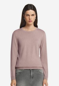 Kvinna med långt mörkt hår som bär silverfärgad hoopörhängen, en mauve långärmad tröja och grå jeans, vänd framåt med ett neutralt uttryck.