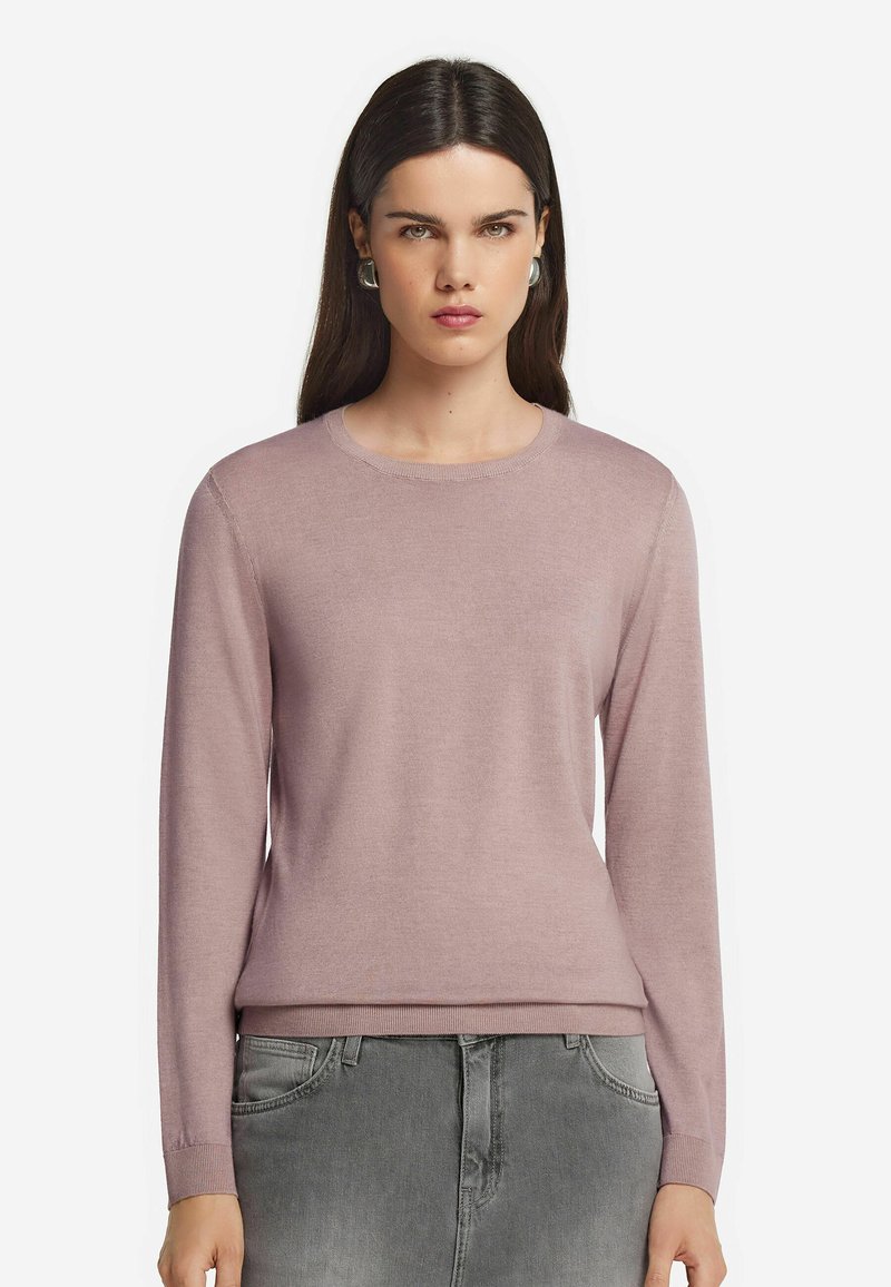 Frau mit langen dunklen Haaren, die silberne Creolen, einen mauvefarbenen Langarmpullover und graue Jeans trägt, nach vorne blickend mit einem neutralen Ausdruck.