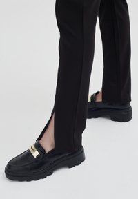 Mocassins noirs avec une semelle épaisse et des accents en métal doré, associés à un pantalon noir ajusté présentant des fentes sur les côtés au niveau de l'ourlet.