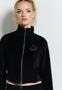 Młoda kobieta z blond włosami, nosząca kolczyki w kształcie kół oraz czarną kurtkę zapinaną na zamek z logo "LH" z kryształków na piersi, na tle jasnej, gładkiej ściany.