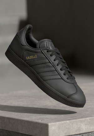 Black Adidas Gazelle sneaker with textured sole and gold lettering, floating above a concrete ledge against a dark gray background.