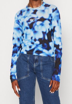 Persona vistiendo una camiseta de manga larga azul y negra con estampado tie-dye con el logo "KARL LAGERFELD JEANS" y jeans de mezclilla azul de cintura alta.