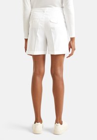 Bílé skládací šortky s ohrnutým lemem, s dvěma zadními kapsami se zapínáním na knoflíky, předvedené z zadní strany, v kombinaci se světlými teniskami.