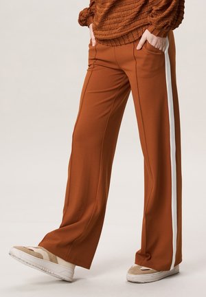 Persoon die rosbruine wijde broek met witte zijstrepen en beige sneakers draagt, staand met handen in de zakken.