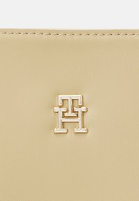 Portefeuille en cuir beige clair avec un emblème logo en ton doré. Texture lisse avec des détails de surpiqûre subtils autour des bords.