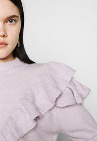 Femme aux yeux bleus portant un pull violet clair avec un volant double couche sur l'épaule et des boucles d'oreilles argentées en forme d'anneau.