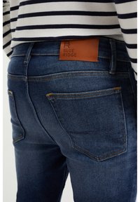 Mörkblå tajta jeans i en strukturerad denimfabric. Har en brun läderlapp med texten "BLUE RIDGE" på bakre midjan och orangea sömmar.