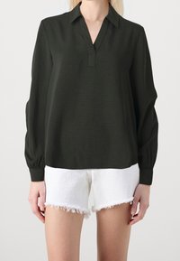 Blouse vert foncé à manches longues et décolleté en V, en tissu texturé. Associée à un short en denim blanc effiloché.