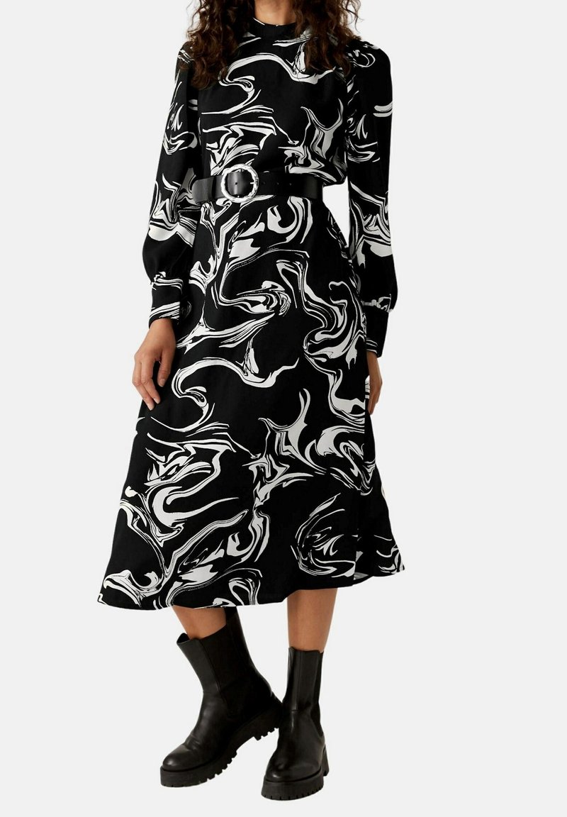 Femme portant une robe midi à manches longues à motif tourbillon noir et blanc avec une ceinture noire et des bottines noires.