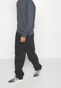 Felpa grigia a girocollo con piccolo logo, abbinata a jeans scuri a vestibilità ampia e sneaker blu con strisce bianche e lacci.