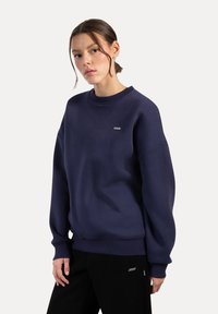 Námořnická modrá mikina vyrobená z měkké tkaniny s kulatým výstřihem, dlouhými rukávy a žebrovanými manžetami. Obsahuje malou značkovou etiketu na hrudi.