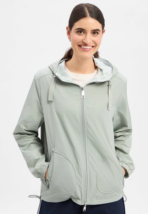 Jonge vrouw die glimlacht, draagt een lichtgrijze zip-up capuchonvest met handen in de zakken tegen een effen witte achtergrond.