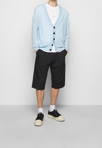 Cardigan lavorato a maglia di colore azzurro chiaro con motivo, bottoni neri, t-shirt bianca, pantaloni corti neri e sneaker nere con suole di colore crema.