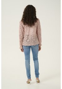 Donna con capelli ricci che indossa una blusa rosa in pizzo a maniche lunghe, jeans skinny azzurri e sandali con tacco bianchi, in piedi con la schiena rivolta in avanti.