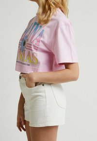 Vaaleanpunainen lyhythihainen t-paita, jossa on monivärinen kukkakuva ja "KANSAS"-teksti, yhdistettynä valkoisiin korkeavyötäröisiin farkkushortseihin.