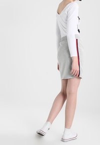 Weißes Langarmoberteil mit rundem Ausschnitt, kombiniert mit einem hellgrauen Rock mit roten Seitenstreifen. Weiße High-Top-Sneaker vervollständigen den Look.