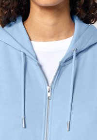 Felpa con zip di colore azzurro chiaro, dal texture morbido, con cordino e dettagli in metallo argentato, indossata sopra una maglietta bianca.