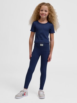 Jong meisje met krullend haar, gekleed in een marineblauw geribd topje met korte mouwen en bijpassende legging met witte sneakers, staand voor een effen achtergrond.