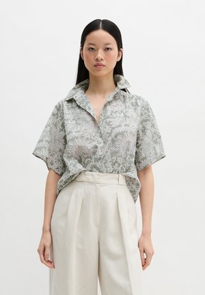 Frau mit langen schwarzen Haaren trägt ein lose sitzendes, kurzärmeliges, gemustertes Hemd, das in hoch taillierte, plissierte, cremefarbene Hosen gesteckt ist.