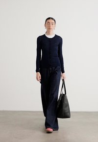 Marinblå ruggad cardigan över en vit topp, ihop med mörk denim vidbyxor. Modellen bär en svart tote-väska och har på sig rosa slip-on skor.