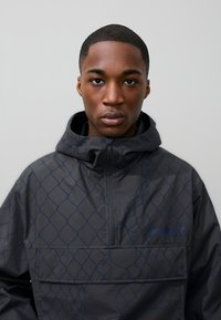 Rövid hajú fiatal férfi fekete kapucnis kabátban, kék láncszemmintával, szemben egy sima háttér előtt.