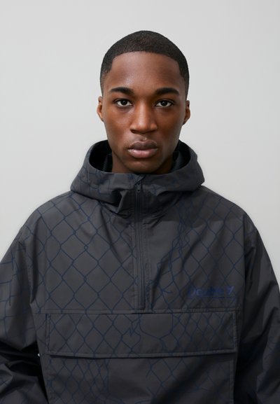 Jeune homme aux cheveux courts portant une veste à capuche noire avec un motif à maillons bleus, faisant face à l'avant sur un fond uni.