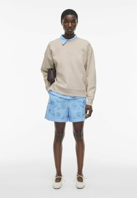 Beżowa bluza ze spadłymi ramionami, jasnoniebieska koszula z kołnierzykiem pod spodem, niebieskie haftowane szorty i kremowe buty z wiązaniem wokół kostki.