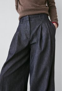 Pantalon en denim foncé avec un devant plissé, une coupe ample et des poches latérales. Tissu texturé et détail de ceinture ajustée. Design décontracté.