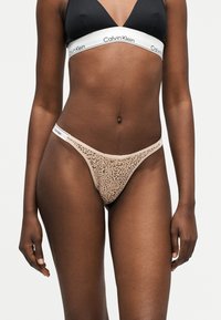 Γυναίκα που φοράει μαύρο τρίγωνο σουτιέν και μπεζ στρινγκ με λεοπάρ σχέδιο, και τα δύο με λευκές ελαστικές λωρίδες με την επωνυμία Calvin Klein.