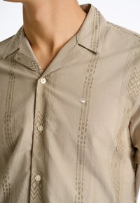 Korte mouwen beige shirt gemaakt van zacht stof, met ingewikkelde lichtbruine patronen en een klein geborduurd embleem op de linkerborst.